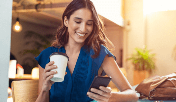 Woman smiling, looking at her phone in one hand with a takeaway coffee in the other.