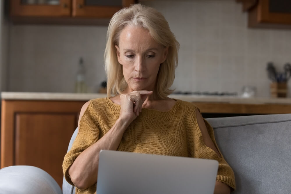 older woman sat thinking looking down at laptop